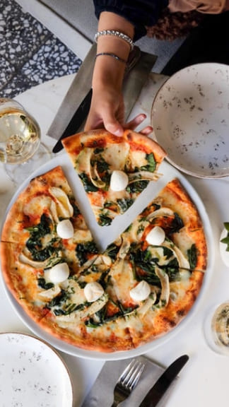 Girl holding one slice of artichoke pizza, the table has a spread of wine and marble dining ware